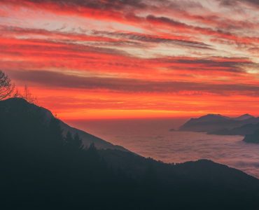 Silhouette of trees on a mountain against a red-streaked cloudy sky, fog and mountains in the distance. Original public domain image from Wikimedia Commons